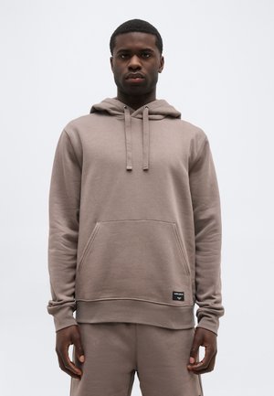 Uomo che indossa una felpa con cappuccio color taupe e pantaloni abbinati, in piedi davanti a uno sfondo grigio chiaro uniforme.