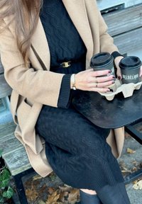 Beige kappa över en svart kabelstickad klänning, som håller två koppar i en kartonghållare på ett svart bord. Släta, texturerade tyger kontrasterar.