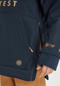 Giacca navy con accenti dorati, dotata di una tasca con zip, polsini elastici e una toppa con logo. Tessuto morbido con una vestibilità comoda.