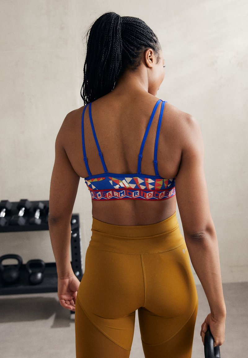 Sujetador deportivo azul con patrones geométricos coloridos y tirantes finos, combinado con leggings marrones de cintura alta, en un fondo de gimnasio.
