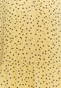 Blouse jaune avec un motif à pois marron foncé, dotée de plis verticaux et d'une fermeture à boutons sur le devant. Tissu doux et texturé.