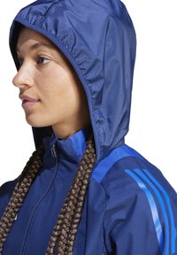 Veste à capuche bleu marine avec une texture brillante, dotée de bandes latérales bleues et d'une fermeture éclair à l'avant. Conçue pour les activités en plein air.