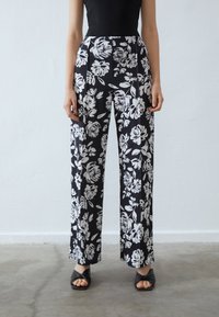 Pantalons noirs à motifs floraux avec des fleurs blanches, design à jambes larges, taille haute et tissu lisse. Associés à des sandales noires à bout ouvert.