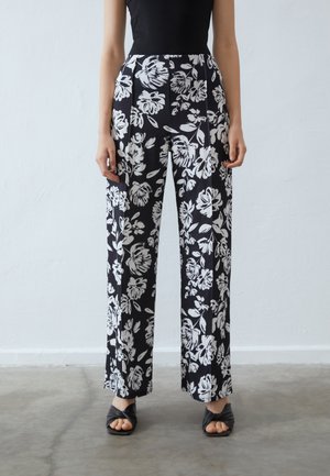 Schwarzblühende Hose mit weißen Blumen, weit geschnittenes Design, hohe Taille und glatter Stoff. Kombiniert mit schwarzen Sandalen mit offenen Zehen.