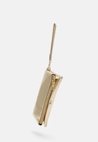 Petit porte-monnaie doré brillant avec poche à fermeture éclair et bandoulière de poignet attachée sur fond clair.