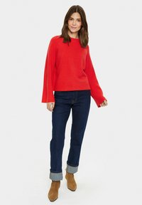 Maglione rosso lavorato a maglia con maniche larghe, abbinato a jeans in denim scuro e stivaletti alla caviglia marroni. Il maglione ha un collo rotondo e una vestibilità comoda.