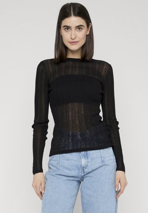 Kobieta o prostych ciemnych włosach, ubrana w przezroczysty czarny top z długimi rękawami na czarnym topie bez ramiączek oraz jasnoniebieskie, wysokie jeansy, stojąca.