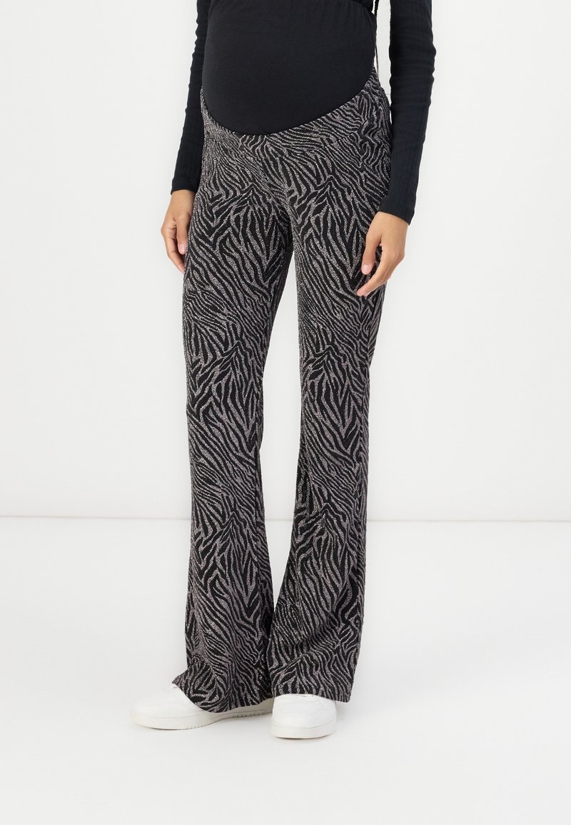 Personne enceinte portant un pantalon de maternité noir et gris imprimé zèbre, associé à un haut noir et des baskets blanches, debout.