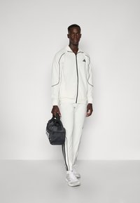 Hvid atletisk tracksuit med sorte accenter, slim fit, og zip-up jakke. Sammen med en grå duffel taske og lysegrå atletiske sko.