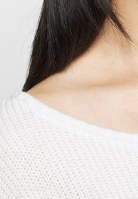 Pull en maille blanche avec un large décolleté, présentant un motif texturé. Le tissu semble doux, avec la lumière se reflétant sur les fibres.