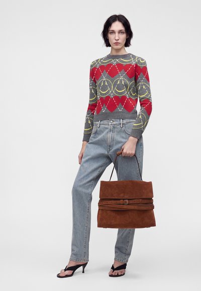Femme portant un pull gris avec des cœurs rouges et des visages souriants jaunes, un jean bleu clair, des sandales noires à talons, tenant un grand sac à main marron.