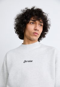 Gray sweatshirt with a high neckline and black embroidered text saying "Service." Features a relaxed fit and soft, textured fabric.