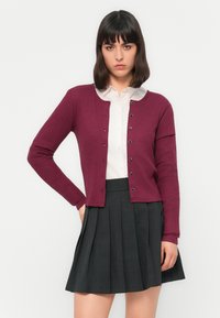 Une femme aux cheveux noirs porte un cardigan en maille bordeaux sur une chemise blanche à boutons et une jupe plissée grise foncée, le tout sur un fond blanc uni.