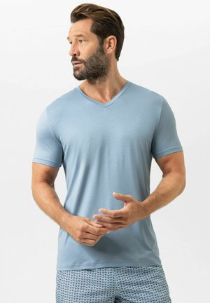 Mann mit Bart, der ein hellblaues T-Shirt mit V-Ausschnitt und gemusterte blaue Shorts trägt, schaut nach links und hat die Hände vor der Taille verschränkt.