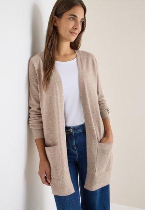 Beige neulecardigan pitkähihaisilla, avonaisella edessä ja kahdella etutaskulla. Pehmeä tekstuuri, rento istuvuus, yhdistetty valkoiseen t-paitaan ja sinisiin farkkuihin.