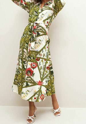 Robe midi à motif floral blanche, avec des accents verts et une ceinture à nouer. Dotée d'un col et de manches longues. Portée avec des talons blancs à brides.