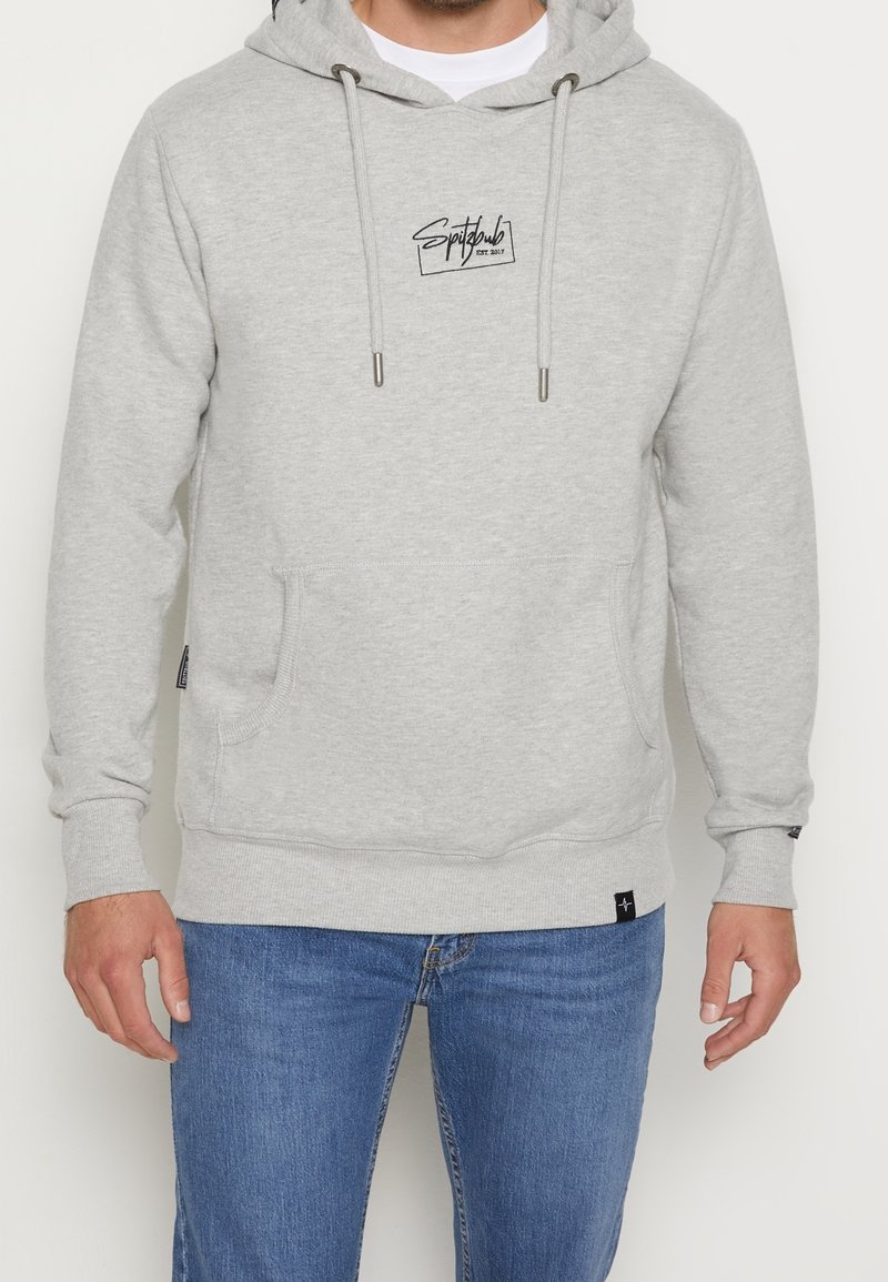 Homme portant un sweat à capuche gris clair avec poche avant et logo "Spitzbub", associé à un jean bleu, debout devant un fond blanc.