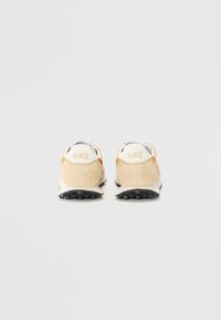 Ett par beigefärgade Nike-sneakers visade från baksidan, med vita hälflikar och svarta strukturerade sulor mot en vit bakgrund.