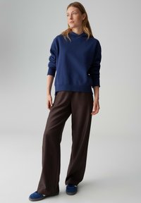 Marineblauer Kapuzenpullover mit langen Ärmeln, kombiniert mit braunen Hosen mit weitem Bein und blauen Schuhen. Vor einem neutralen Hintergrund fotografiert.