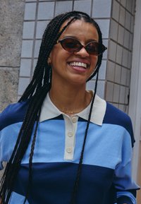 Šypsanti moteris su ilgais kaspinais, vilkinti ovaliais saulės akiniais ir mėlynos dryžuotos polo marškinėliais, stovi lauke priešais plytelių sieną.