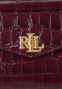 Burgundi színű műgyík bőr kézitáska arany színű díszítéssel és zárható csattal, a táska elején pedig egy kiemelkedő monogram logo található.