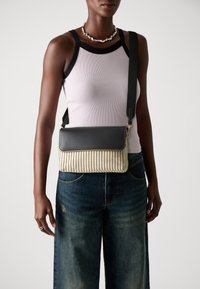 A crossbody táska fekete bőr fedéllel rendelkezik, szőtt bézs és fehér anyagból készült, rácsos mintával, állítható pánttal és aranyszínű fém díszítéssel.