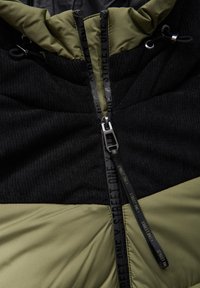 Svart och olivgrön jacka med en strukturerad stickad övre sektion, slät nedre sektion och en central dragkedja med märkta dragtaggar.