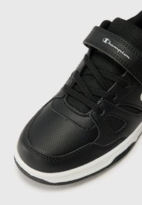 Chaussure de sport noire avec une tige en cuir perforé, une languette en tissu texturé, des lacets et une semelle intermédiaire blanche robuste. Logo de la marque sur la sangle.