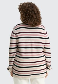 Persoon met krullend haar draagt een gestreepte lange mouwen shirt in wit, roze, bordeauxrood en zwart, staand met de rug naar de camera tegen een lichte achtergrond.