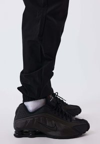 Gambe che indossano pantaloni neri, calze bianche e sneakers Nike Shox nere con molle ammortizzanti visibili su uno sfondo bianco.