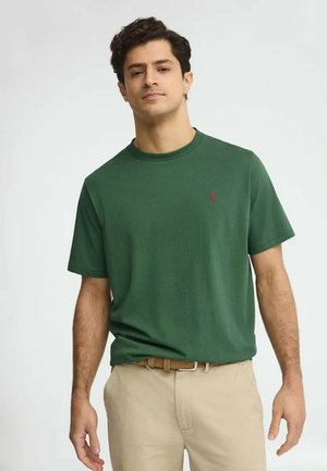 Hombre con camiseta verde de manga corta con logo rojo, pantalones beige y cinturón trenzado, posando frente a un fondo claro y liso.