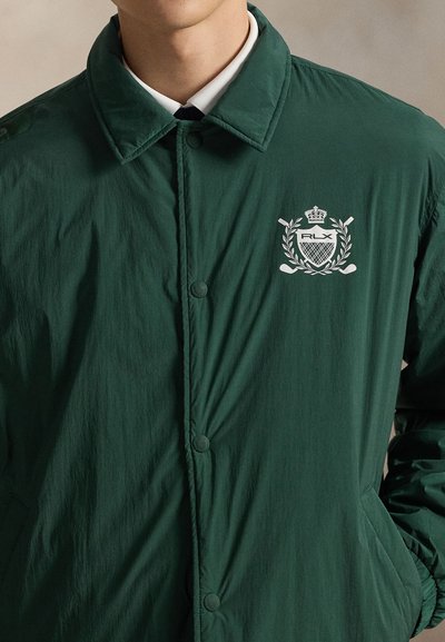 Mees, kes kannab tumerohelist nööbitavat jopet, millel on vasakul rinnal valge RLX vapp, millel on laurelipärg, kroon ja ristatud golfikepid.