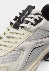 Κοντινό πλάνο γκρι και λευκού αθλητικού παπουτσιού Calvin Klein με υφασμάτινη υφή, λευκά κορδόνια, μαύρη λουρίδα για κράτημα και λογότυπο στο πλάι και στη γλώσσα.