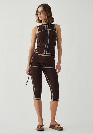 Nő fekete ujjatlan felsőben és világoskék varrású rövid leggingsben, napszemüvegben és barna szandálban, egyszerű háttér előtt áll.