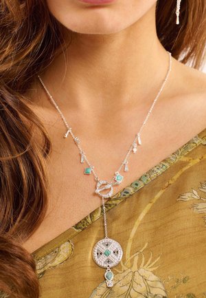 Collier en argent avec un pendentif rond au design géométrique, accentué de cristaux verts et de charms en cascade, sur une fine chaînettes.