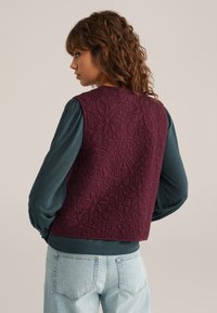 Gilet trapuntato bordeaux con motivo floreale, indossato sopra un top a maniche lunghe teal, abbinato a jeans blu chiaro. Superficie testurizzata con dettagli di cucitura.