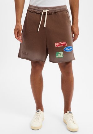 Mann trägt braune Shorts mit Kordelzug und Applikationen, kombiniert mit weißen Turnschuhen und einem weißen Oberteil, steht vor weißem Hintergrund.