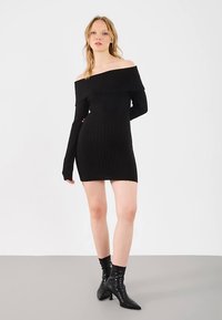 Jauna moteris stovi su viena ranka už nugaros, vilkėdama juodą off-shoulder raštuotą mini suknelę ir juodus aukštakulnius aulinukus.