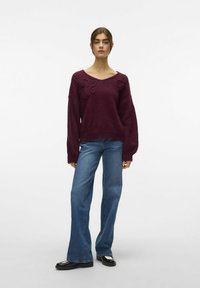 Bordeaux gebreide sweater met brede mouwen en driedimensionale bloemdetails, gecombineerd met uitlopende blauwe jeans en zwarte schoenen.