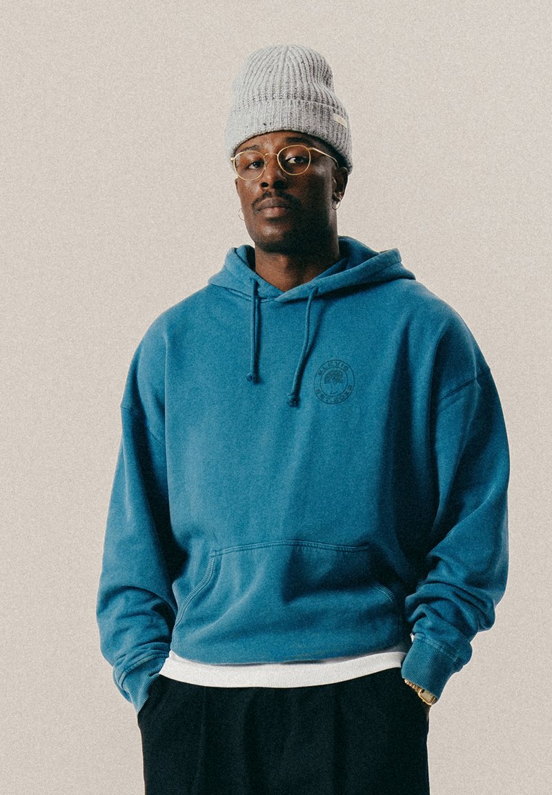 Mężczyzna w szarej dzianinowej czapce, okrągłych okularach, niebieskiej bluzie z kieszenią z przodu, białej koszulce oraz czarnych spodniach, stojący na tle jednolitego tła.