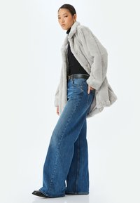 Abrigo de piel sintética gris con un cuello alto, combinado con unos vaqueros de mezclilla azul de pierna ancha. La chaqueta presenta una textura suave y detalles de botones.