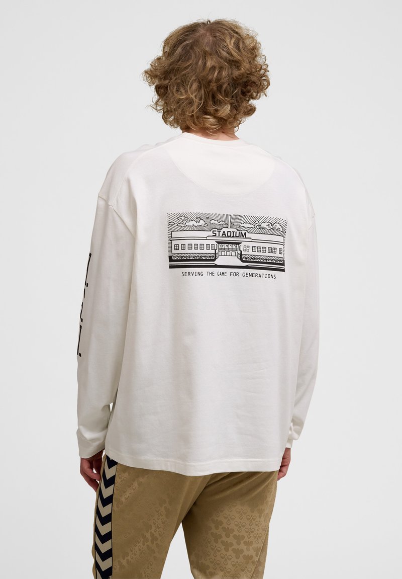 Witte lange mouw shirt met een bedrukte grafiek van een stadion op de achterkant, met de tekst "DEDICATED AAN HET SPEL VOOR GENERATIES" eronder.