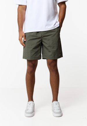 Mężczyzna w oliwkowozielonych szortach, białych trampkach i białej koszulce z krótkim rękawem, z rękami w kieszeniach, stojący na tle jednolitego białego tła.