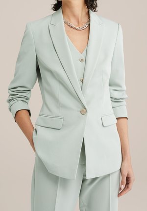 Vrouw draagt lichtgroene, op maat gemaakte blazer en bijpassende broek, met opgerolde mouwen en een dikke zilveren kettink.