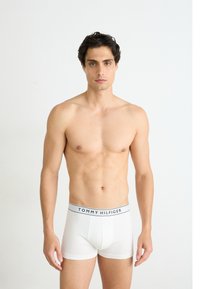 Fehér pamut boxeralsó fekete csíkozott derékkal, amelyen a "TOMMY HILFIGER" logó található; a modell ing nélkül áll egy világos háttér előtt.