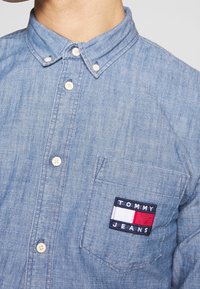 Camicia di jeans azzurro chiaro con colletto button-down, bottoni bianchi, tasca sul petto con una toppa blu navy con il logo "TOMMY JEANS" in rosso e bianco.