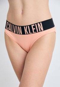 Perzikkleurige bikini slips met een zwarte elastische tailleband waarop het "CALVIN KLEIN" logo in het roze is gedrukt. Vloeiende textuur, low-rise ontwerp.