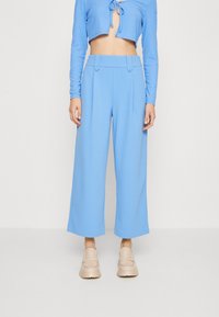 Jasnoniebieskie spodnie w szerokim kroju z wysokim stanem, z dwoma guzikami jako akcentami. Wykonane z gładkiego materiału, połączone z beżowymi butami wsuwanymi.
