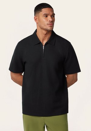 Mann mit kurzen Haaren, der ein schwarzes, kurzärmeliges Polohemd mit Reißverschluss und olivgrüne Hose trägt, steht mit den Händen hinter dem Rücken.