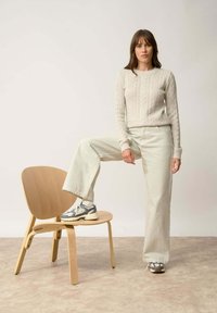 Jasno beżowy sweter w warkocz, bladozielone spodnie w szerokiej nogawce i szare buty sportowe. Drewniane krzesło jest ustawione pod stopą modelki.